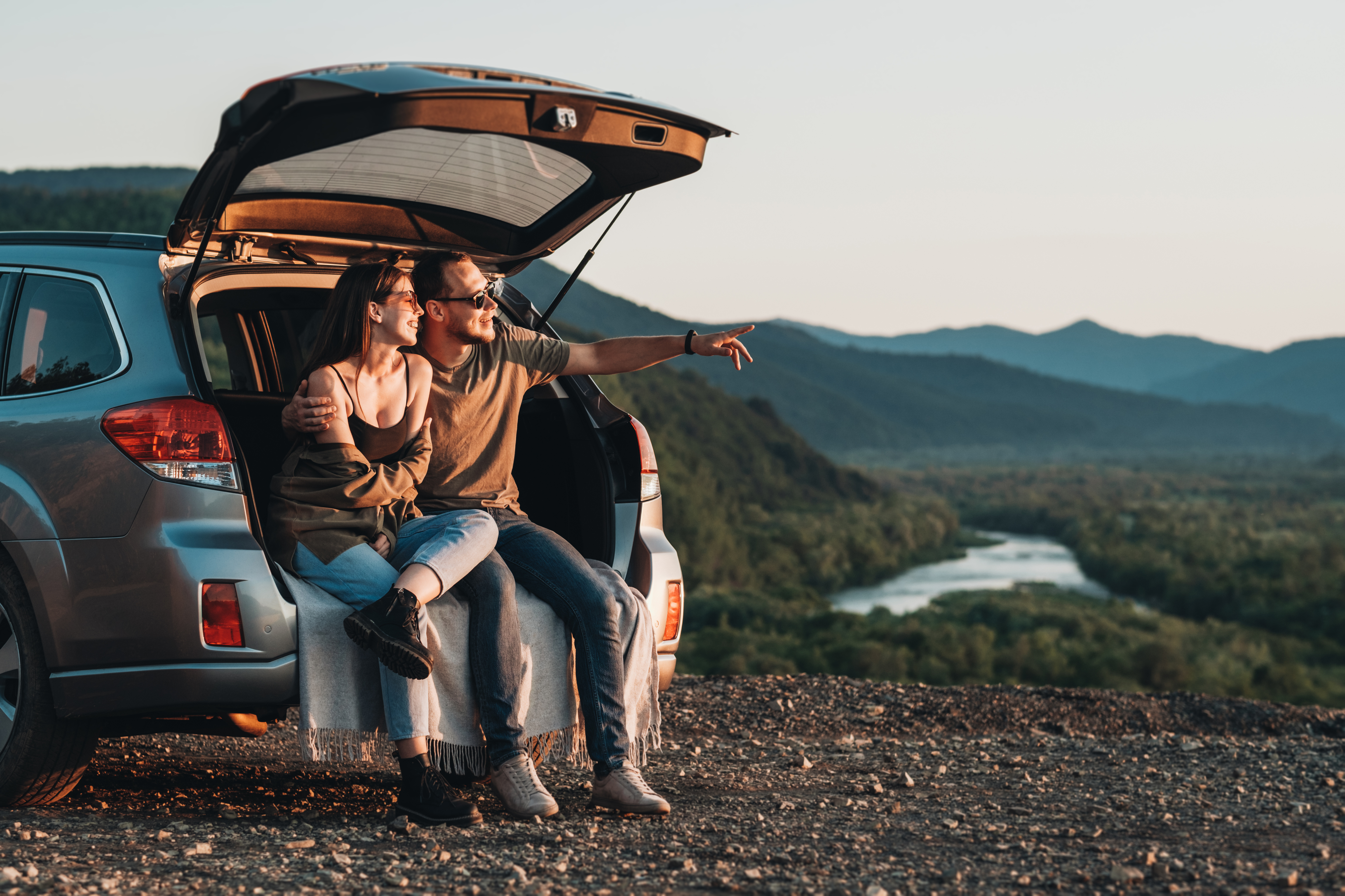 Couple sitting in the open boot of a car overlooking a river valley at sunset during a road trip stop